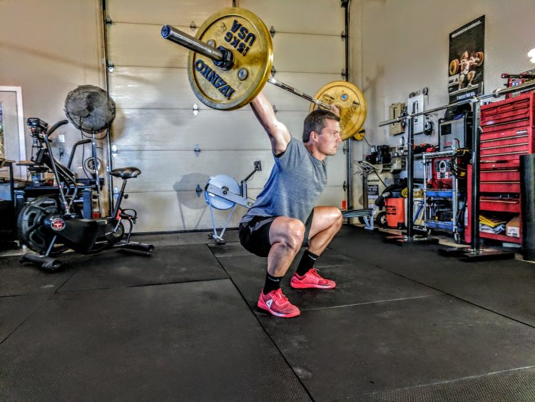 Home gym in two-car garage with man wearing shorts, T shirt and red athletic shoes lifting barbell