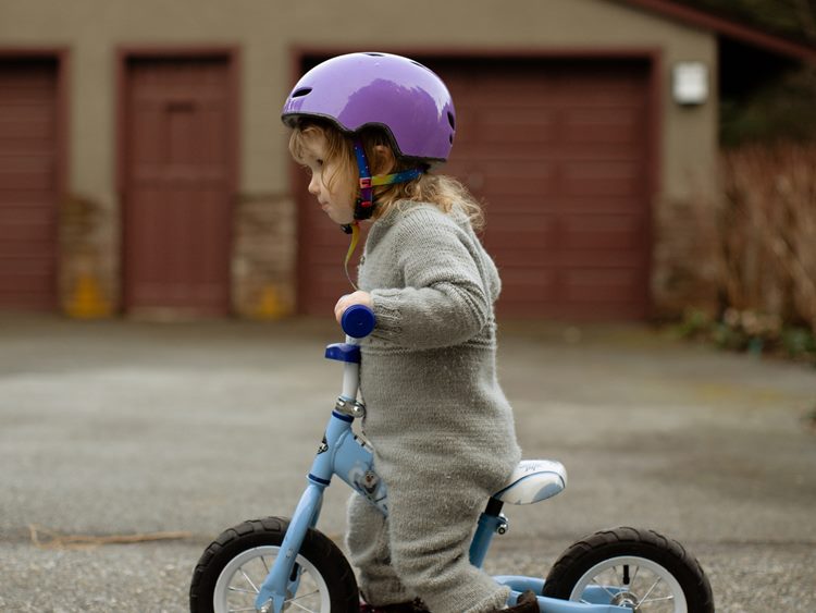 Young girl in purple bike helmet riding small bicycle with driveway and wooden garage doors in background